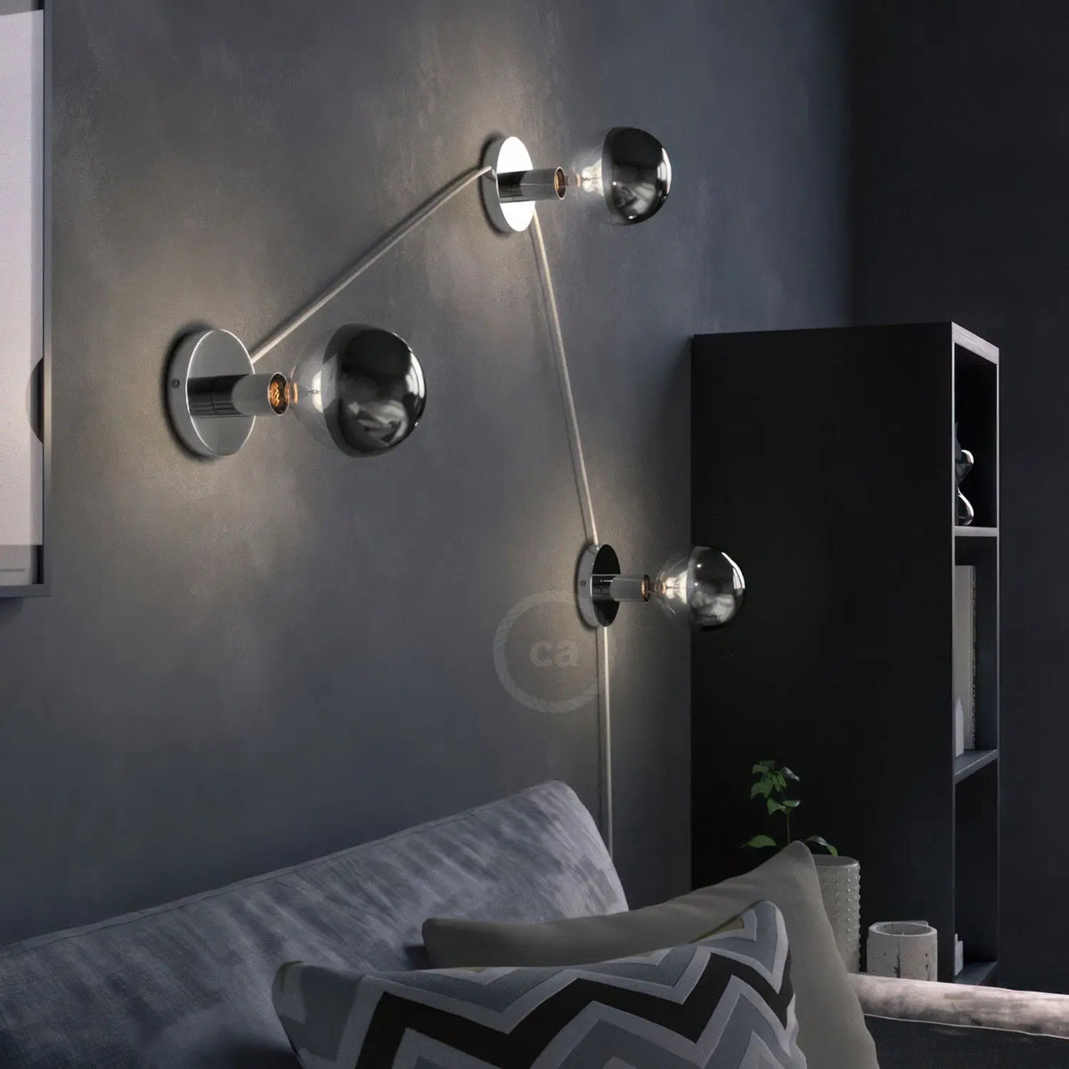 A modern wall-mounted light fixture with three adjustable metallic arms and spherical shades, shown lit in a dark living room setting above a sofa.